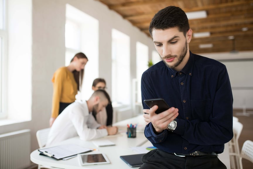 Young business man with beard in dark shirt thoughtfully using cellphone in office with colleagues on background