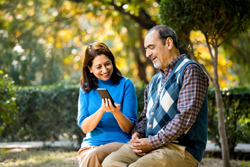 Smiling middle-aged woman showing smartphone to older man in park, symbolizing digital inclusion and tech access for seniors in public spaces