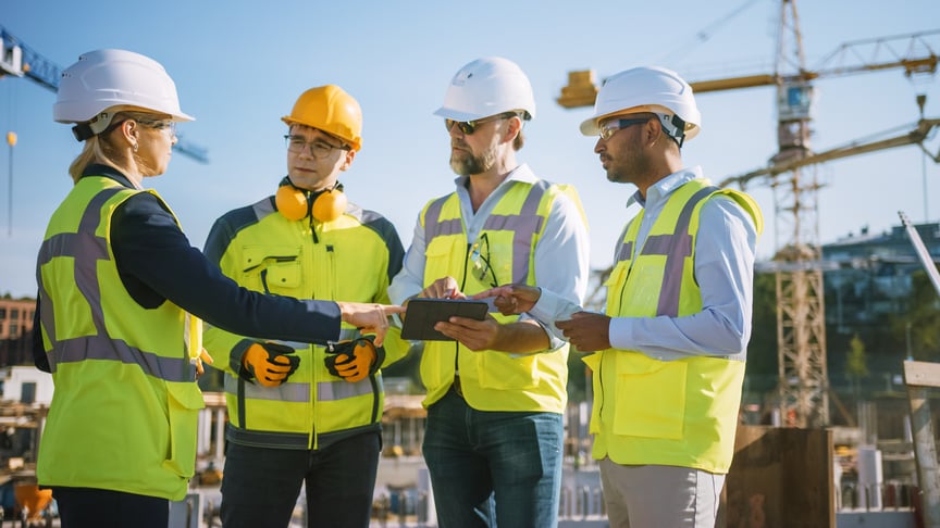 Construction project managers and engineers wearing safety vests and helmets review plans on a tablet at an active construction site under clear blue skies