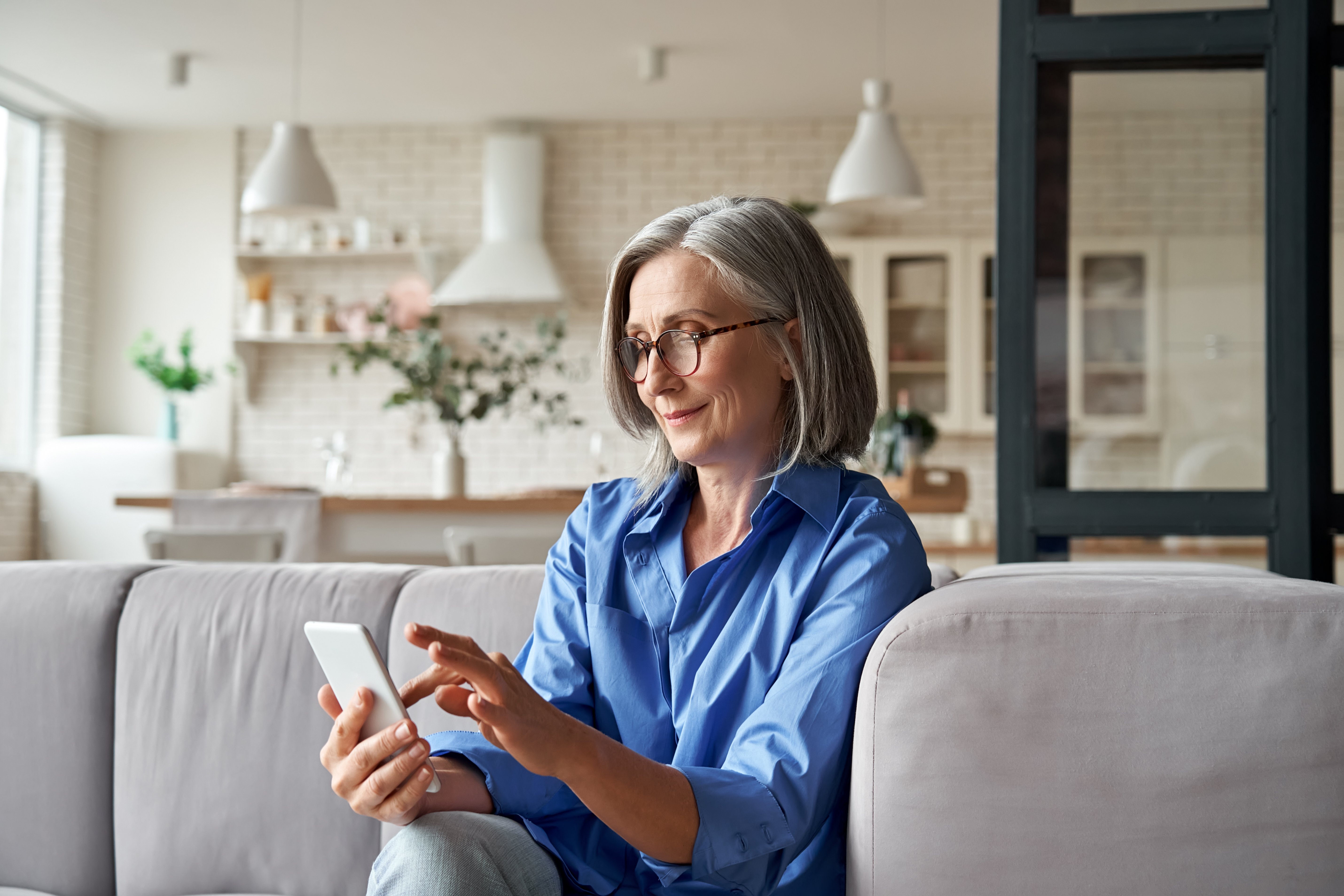 Smiling older woman with gray hair using smartphone while sitting on couch at home, representing senior digital engagement and tech literacy
