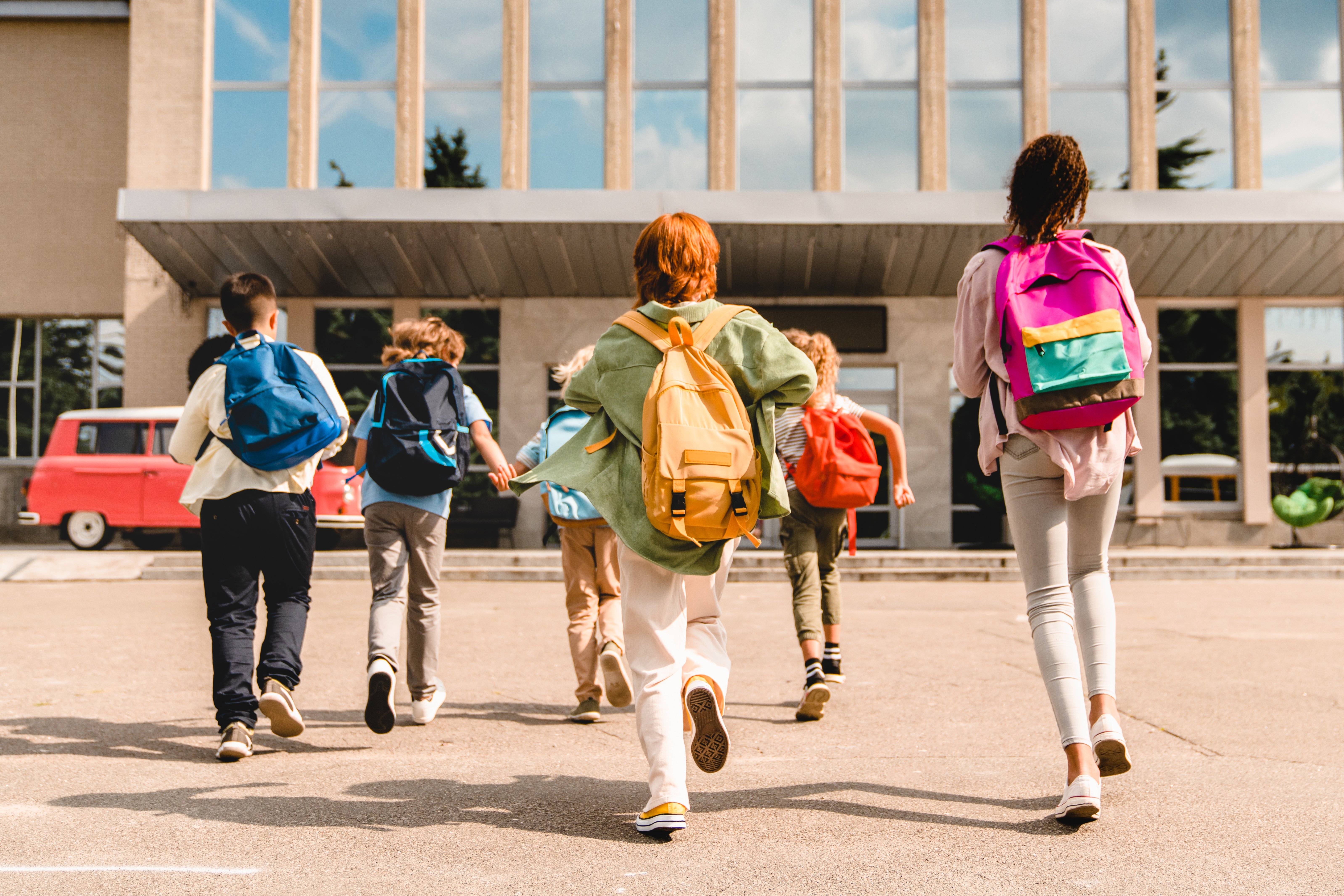 Group of schoolchildren wearing colorful backpacks running toward school building on a sunny day, excited for the new school year