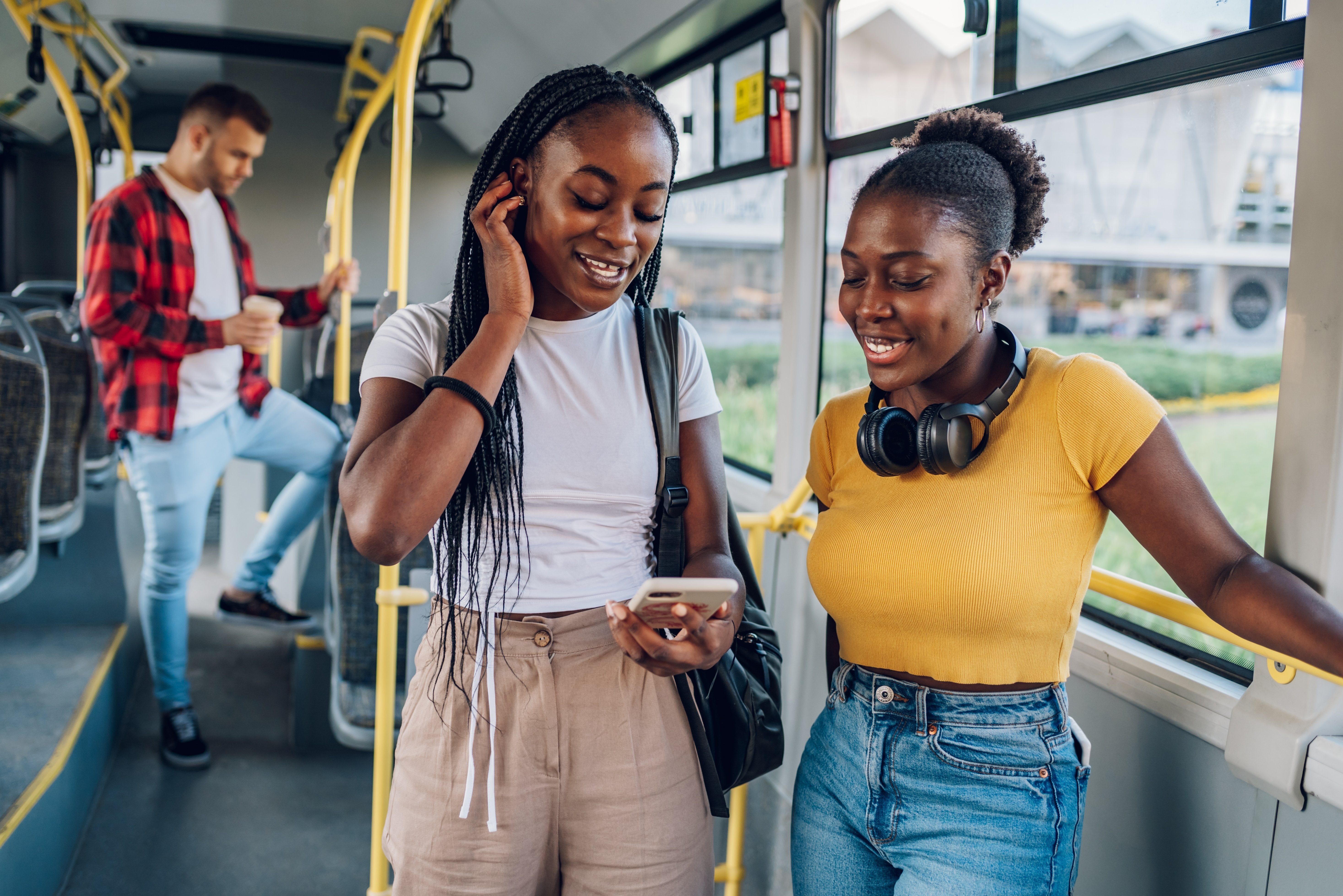 Two young women smiling and looking at a smartphone while riding a city bus, enjoying music and conversation during their commute