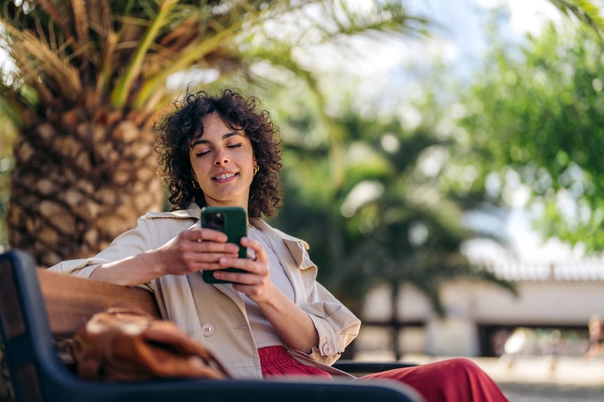 Young woman sitting on a park bench using smartphone, enjoying a sunny day surrounded by palm trees