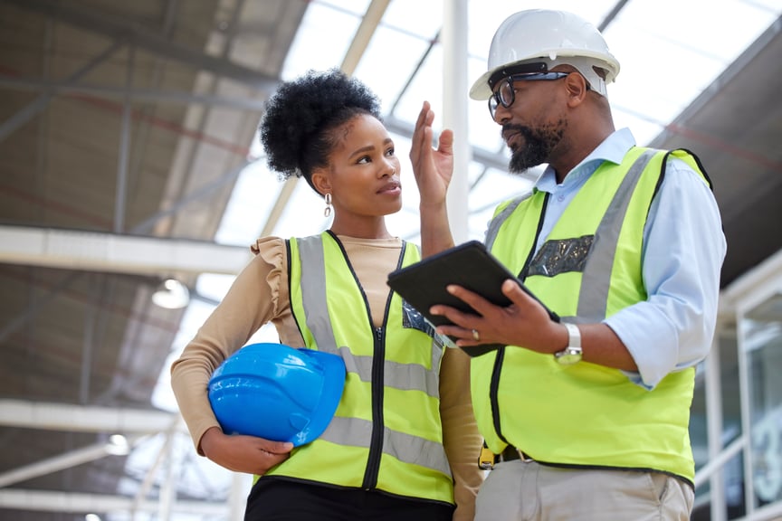 Male and female engineers in safety vests discussing project details using publiq app inside an industrial building; woman holding blue hard hat