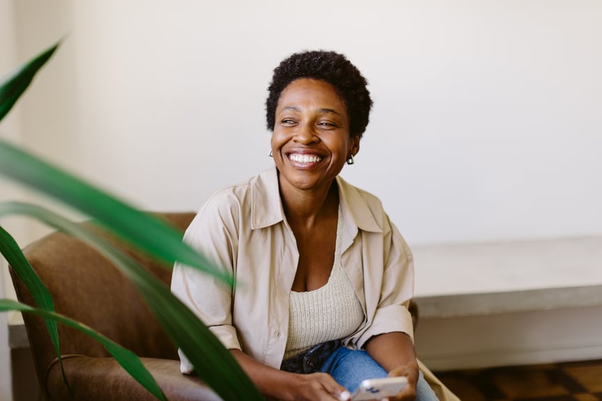 Smiling woman sitting indoors with a smartphone in hand, enjoying a relaxed and positive moment