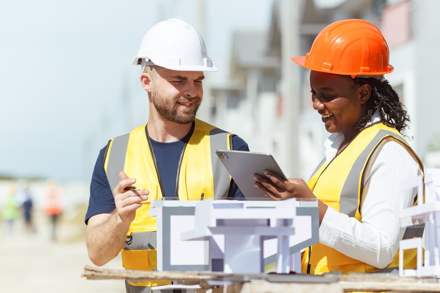 Male and female construction workers wearing hard hats and safety vests reviewing building plans on a tablet at a construction site