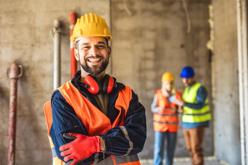 Smiling construction worker wearing a hard hat and safety vest at a job site with coworkers discussing plans in the background