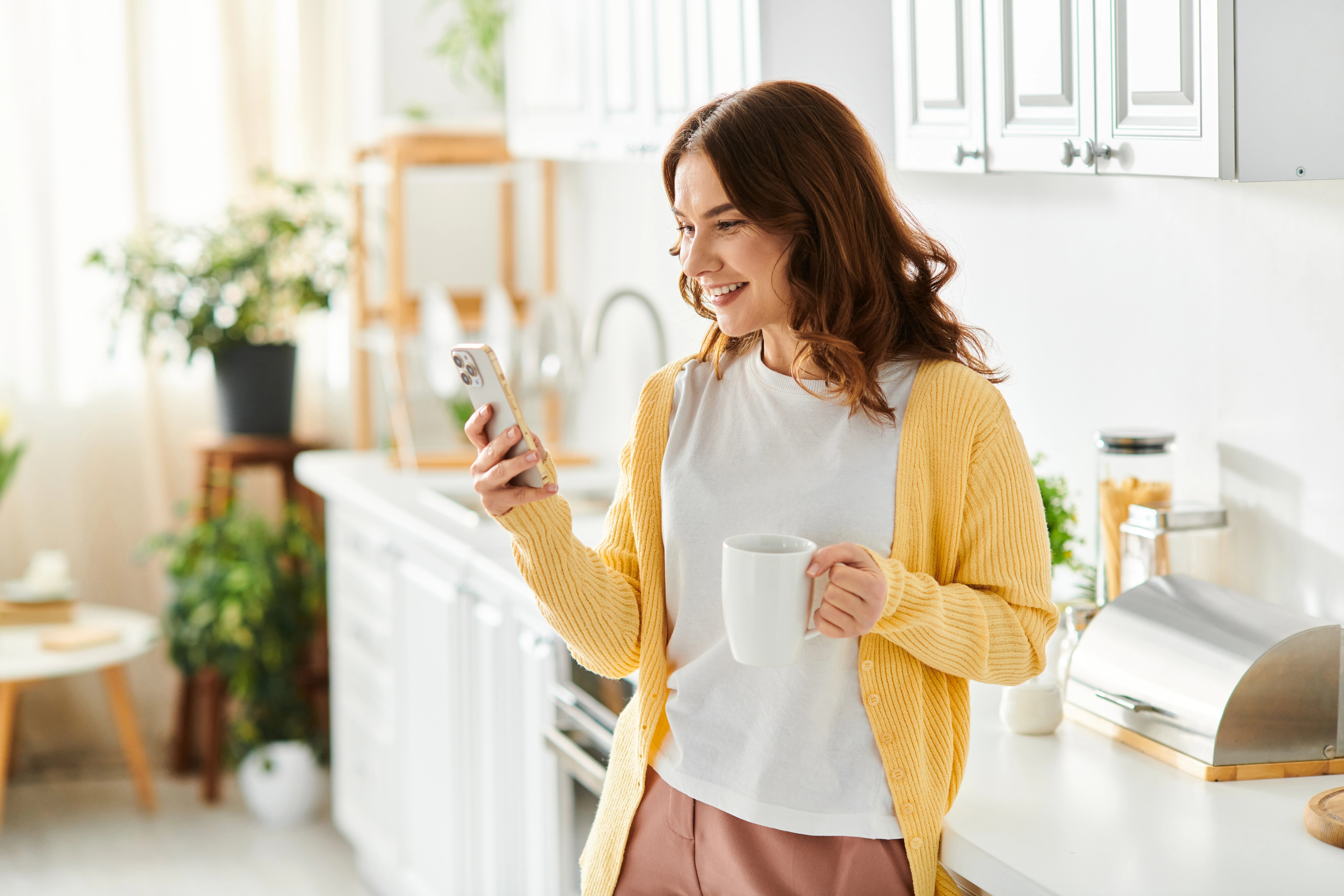 Happy young woman in cozy kitchen holding a mug and smiling at her smartphone, enjoying a relaxed morning at home
