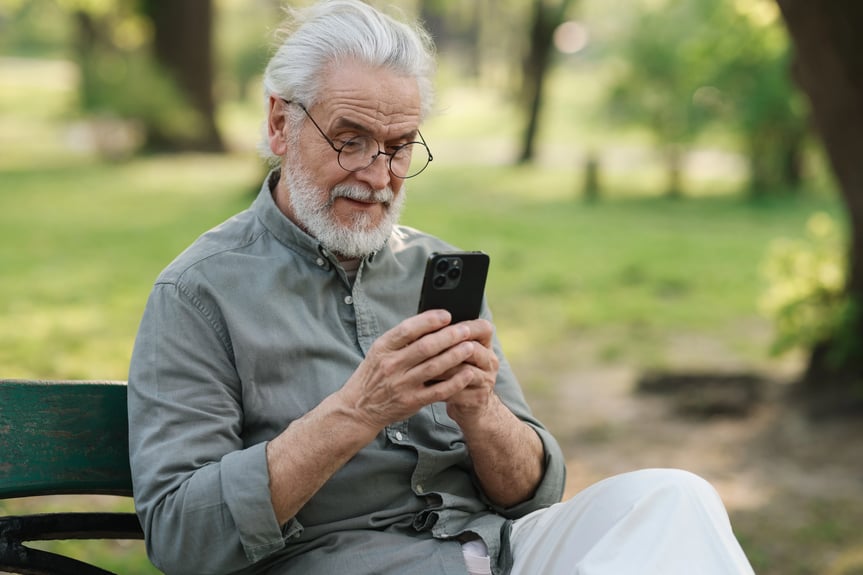 Elderly man sitting on a park bench using his smartphone and smiling while enjoying time outdoors