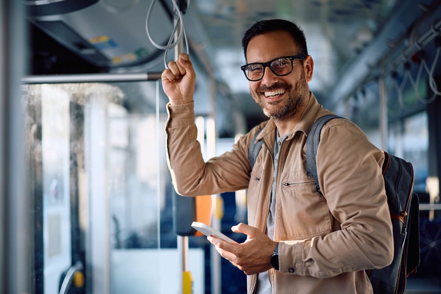 Smiling man wearing glasses and a backpack holding a smartphone while standing on a bus during his commute