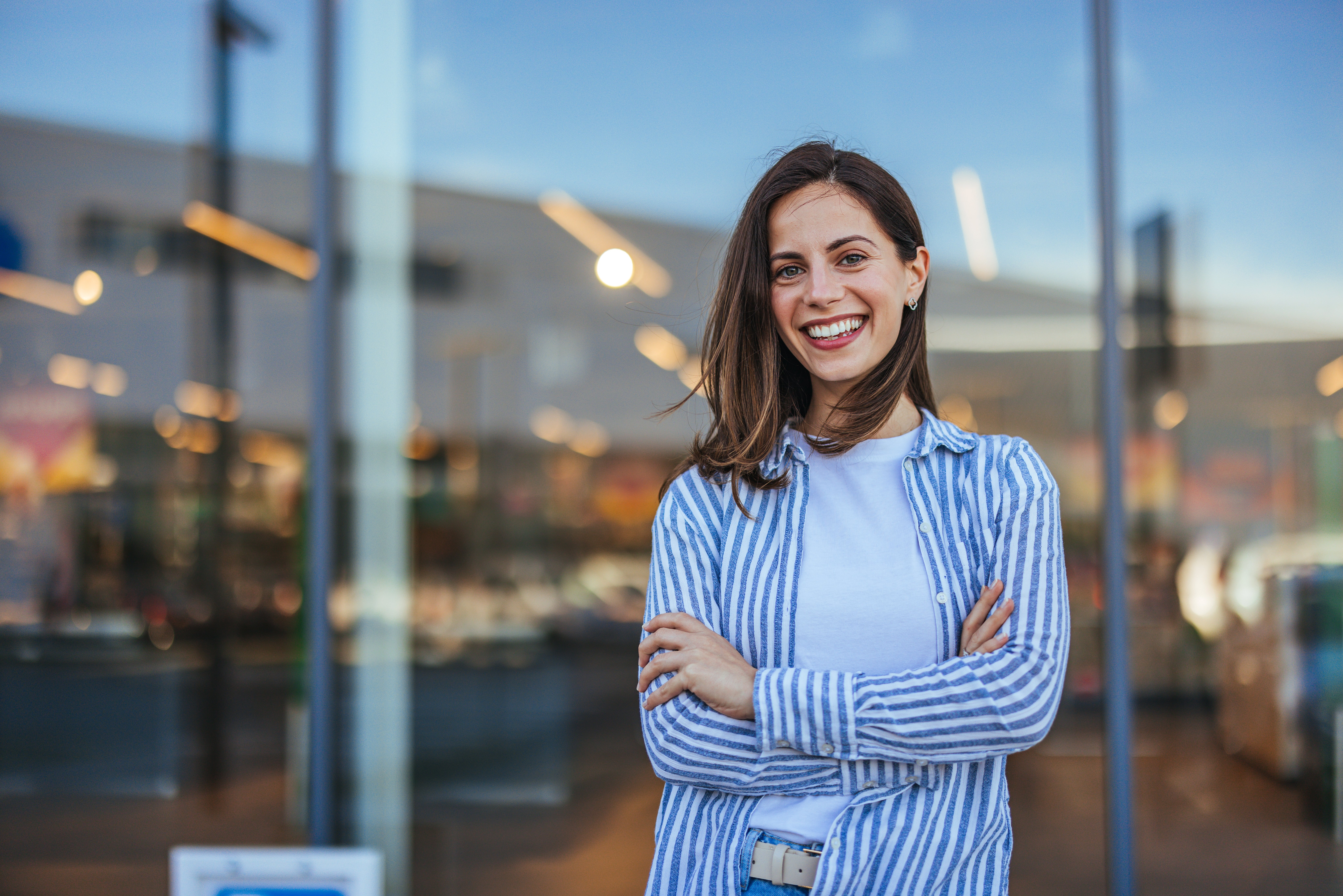 Confident young woman smiling with arms crossed outside a modern glass-fronted building, wearing casual striped shirt