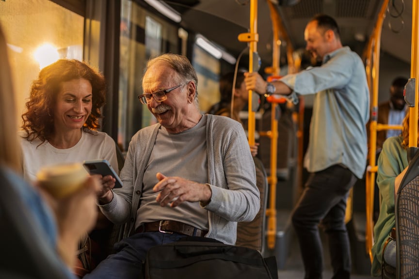Happy older man and woman using smartphone together on a public bus, surrounded by passengers during commute