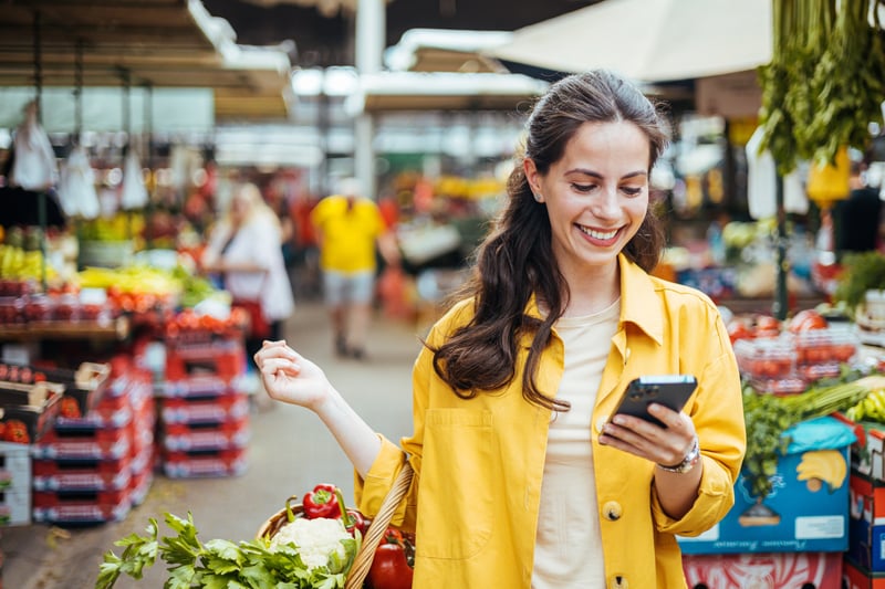 Smiling woman holding a smartphone while shopping for fresh produce at an outdoor market