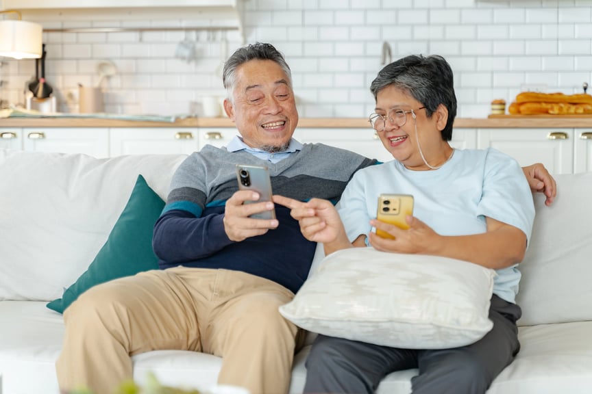 Happy senior couple sitting on a sofa at home, sharing something on their smartphones and smiling together