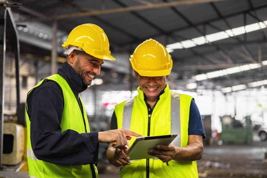 Two smiling factory workers in safety vests and yellow hard hats reviewing information on a digital tablet inside an industrial facility