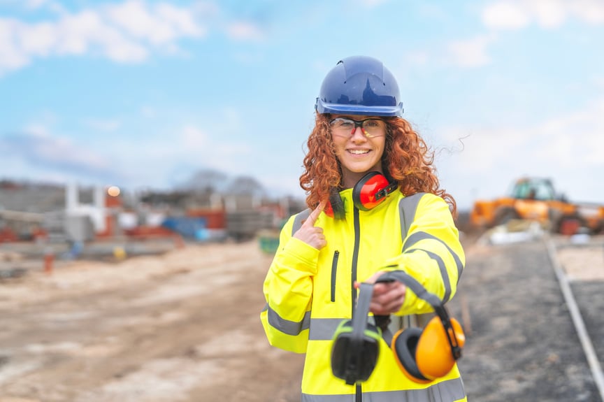 Female construction worker wearing a high-visibility jacket and hard hat, smiling and holding safety earmuffs at a worksite