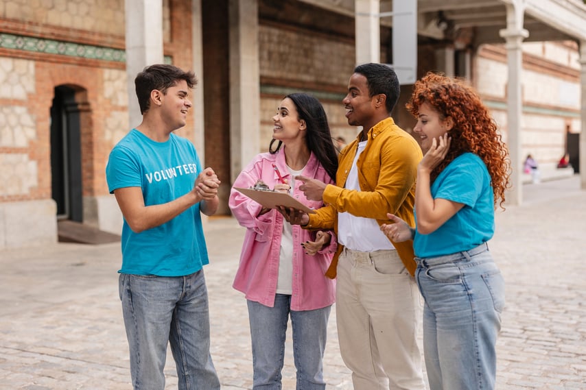 Group of young volunteers standing outdoors, smiling and discussing plans while holding a clipboard during a community project.