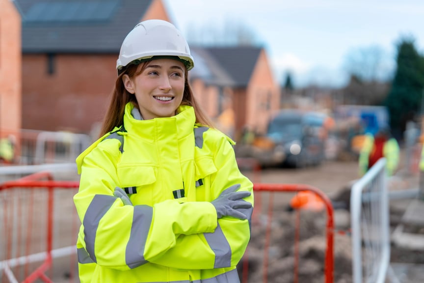 Female construction worker wearing a high-visibility jacket and hard hat, smiling confidently at a housing development site