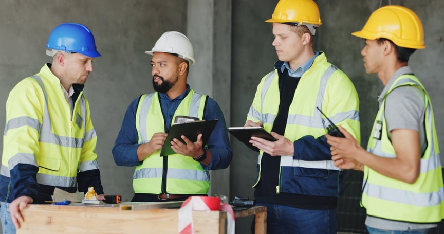 Group of construction workers and engineers in safety vests and hard hats discussing plans on a job site with digital tablets and clipboard
