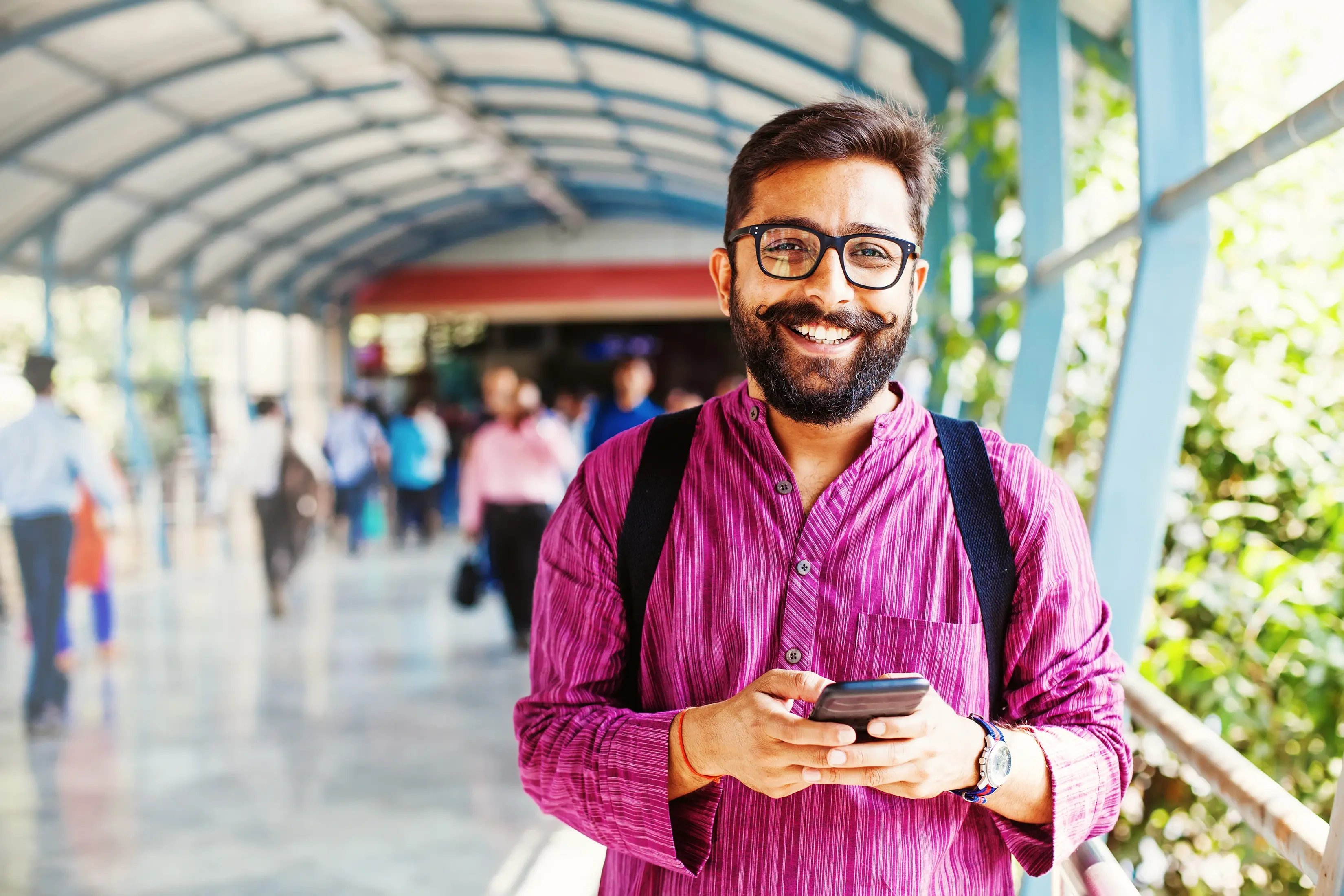 Smiling young man in glasses and traditional kurta using smartphone at a train station walkway, with commuters in the background
